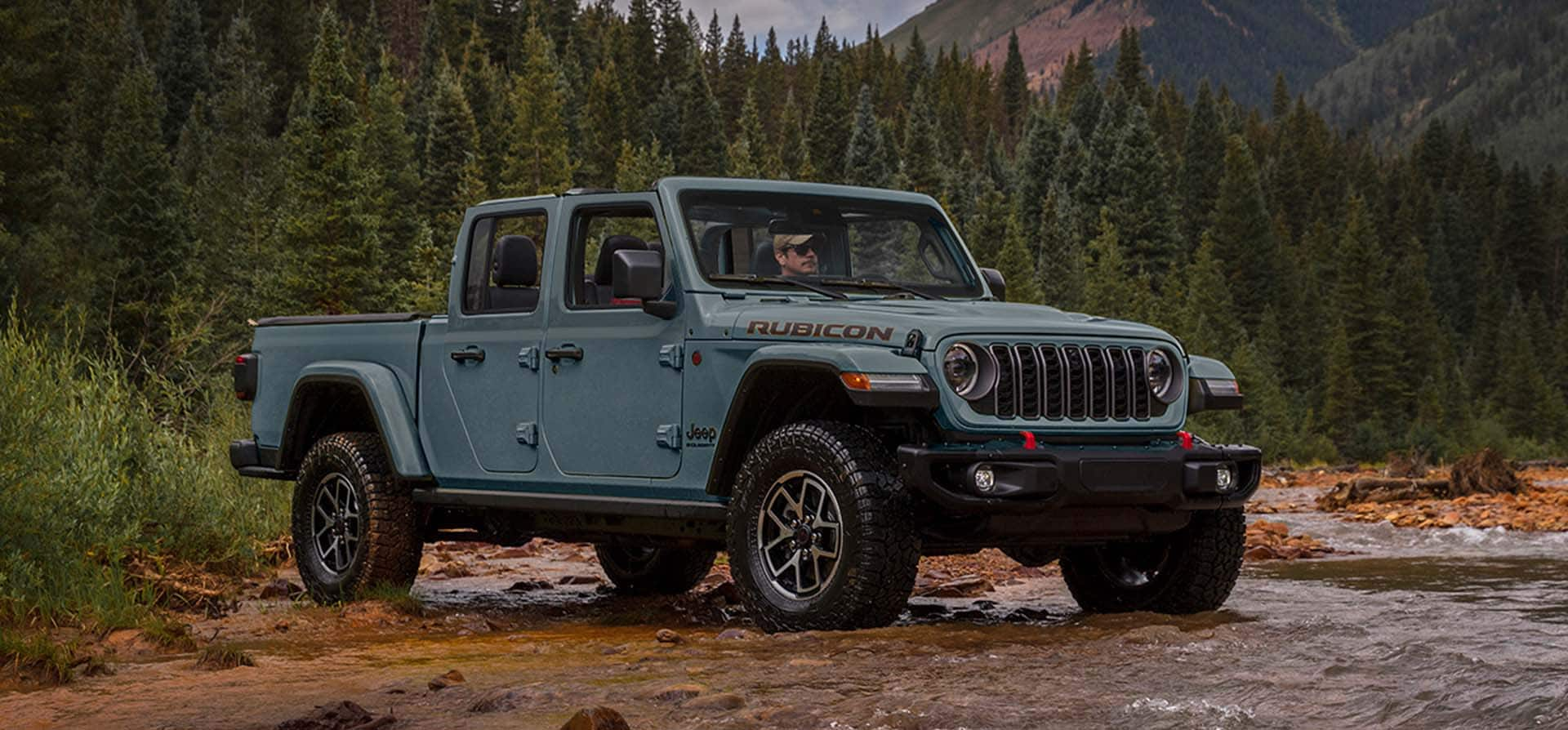 A blue 2025 Jeep Gladiator about to drive through a creek outside a forest.